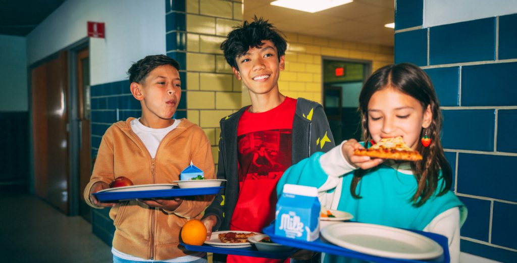 Three children standing in a cafeteria holding lunch trays with pizza and milk. 