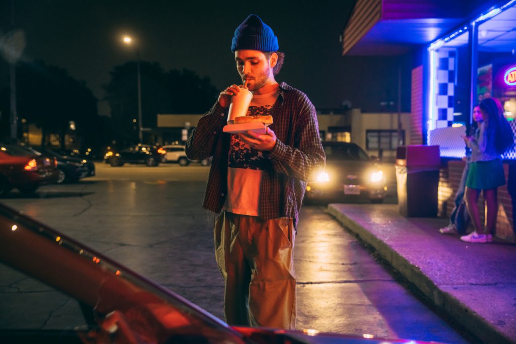 Young man with a beard wearing a blue stocking hat, flannel shirt over a t-shirt walking out of a convenience store at night sipping a soft drink and carrying a slice of to go pizza.  