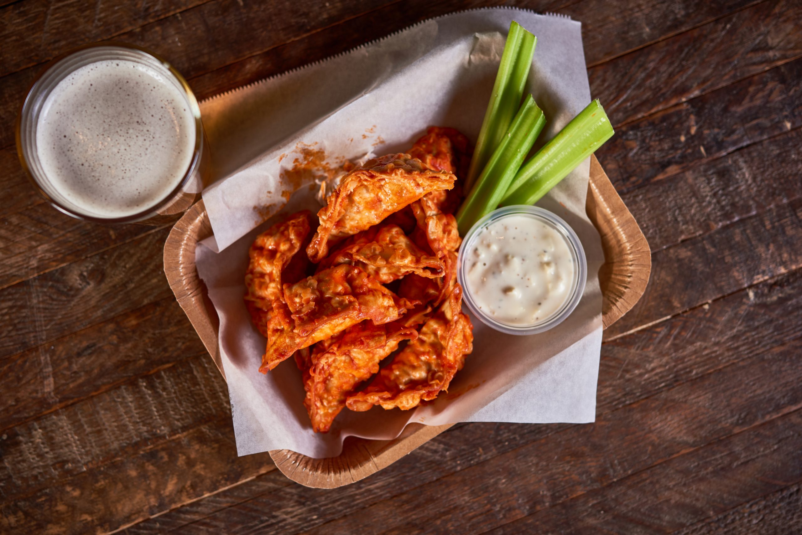 Overhead shot of fried dumplings tossed in buffalo sauce with sides of three celery sticks and a ramekin of blue cheese dressing on a small wooden tray. 