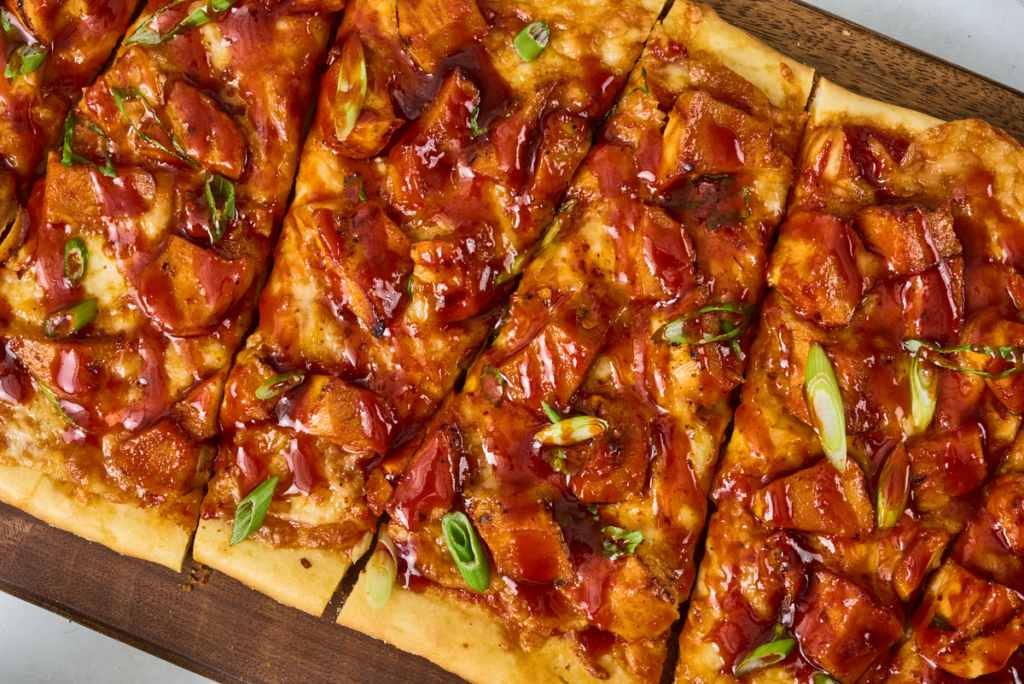 Flatbread pizza topped with chicken, melted cheese, and green onions, sliced into squares on a wooden board.