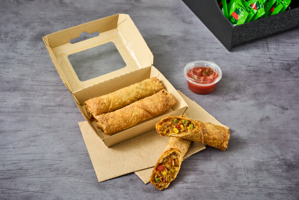 Three MINH® beef taco egg rolls served in a open cardboard takeout container alongside dipping sauce.