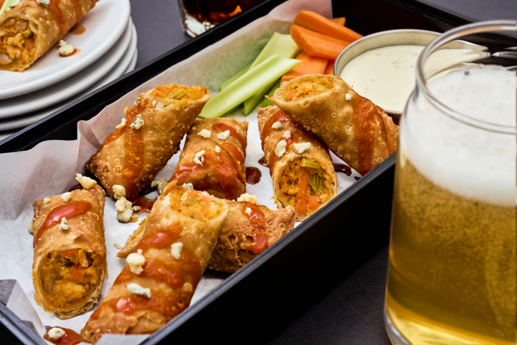 Halved Buffalo chicken egg rolls served on a tray with celery sticks, carrots, and dipping sauce.