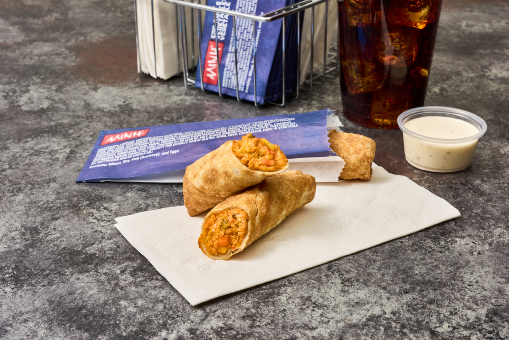 Halved MINH® Buffalo chicken egg rolls served on parchment paper on a convenience store counter next to dipping sauce.