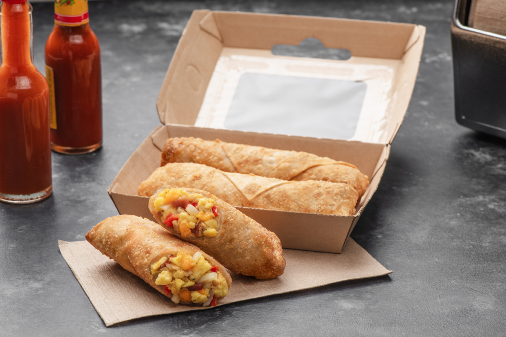 Halved MINH® breakfast egg rolls served in a cardboard take-out container next to dipping sauces.