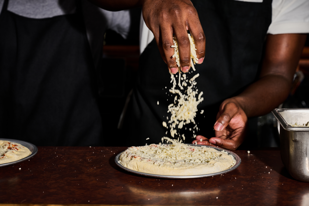 A chef sprinkling grated cheese onto a pizza dough crust on a kitchen counter.